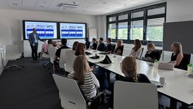A large group of people are sitting in a meeting room looking at two monitors and a man at the front of the room giving a lecture.