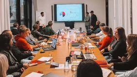 A group of people sitting around a large conference table and follow the presentation of three people at the end of the table.