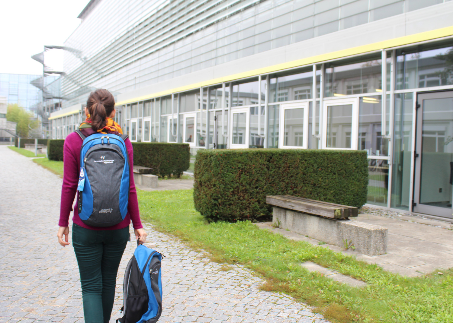 Eine Studentin geht an THI-Gebäuden entlang, auf dem Rücken und in der Hand trägt sie je einen schwarz-blauen THI-Rucksack
