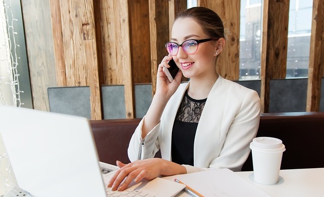 Eine junge Frau im Business-Look sitzt vor ihrem Laptop und telefoniert nebenbei am Handy